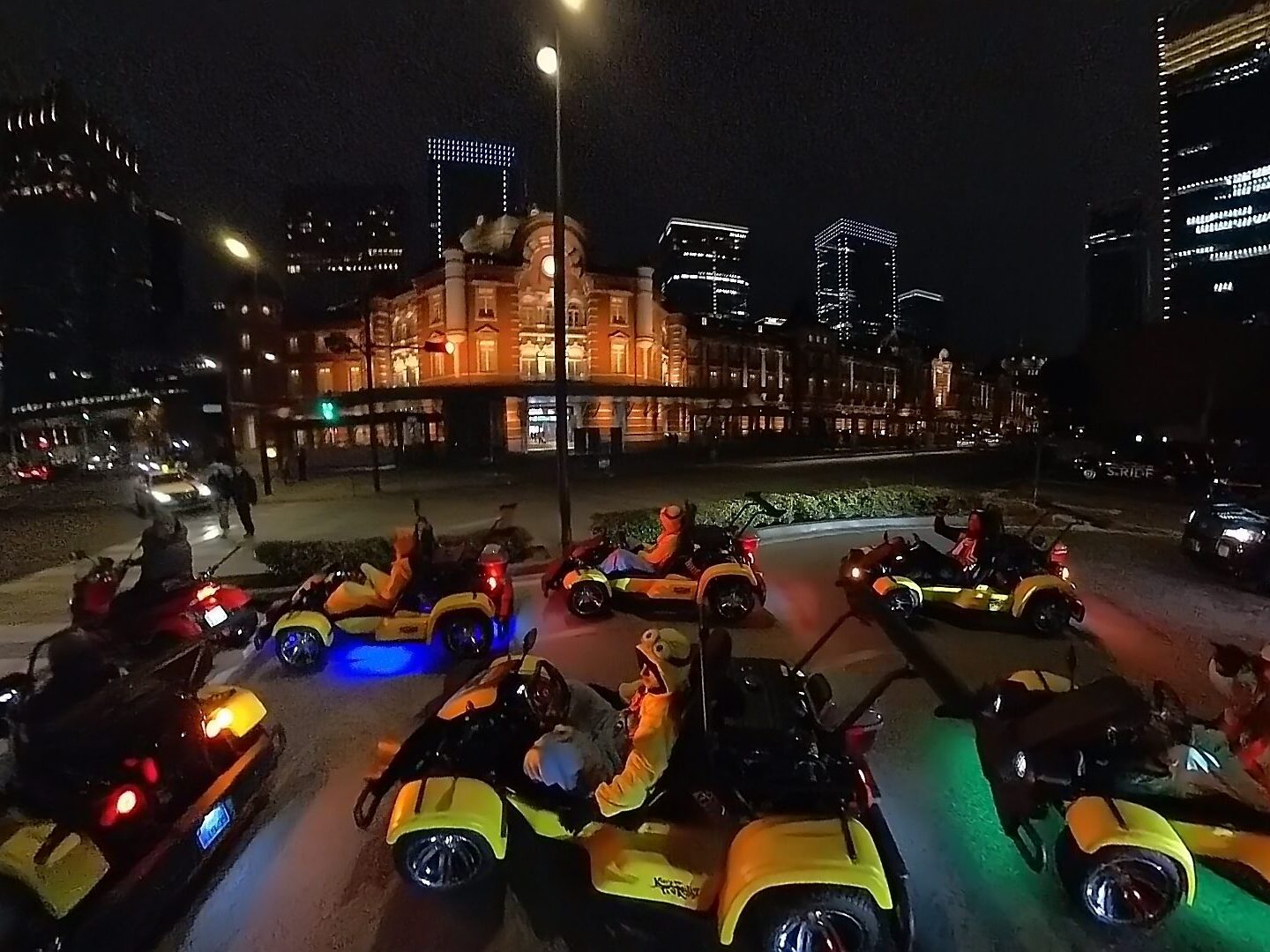 A fleet of bright yellow Kartzilla Go Karts driving in front of the illuminated red brick building of Tokyo Station at night, with partially lit skyscrapers towering in the background.
