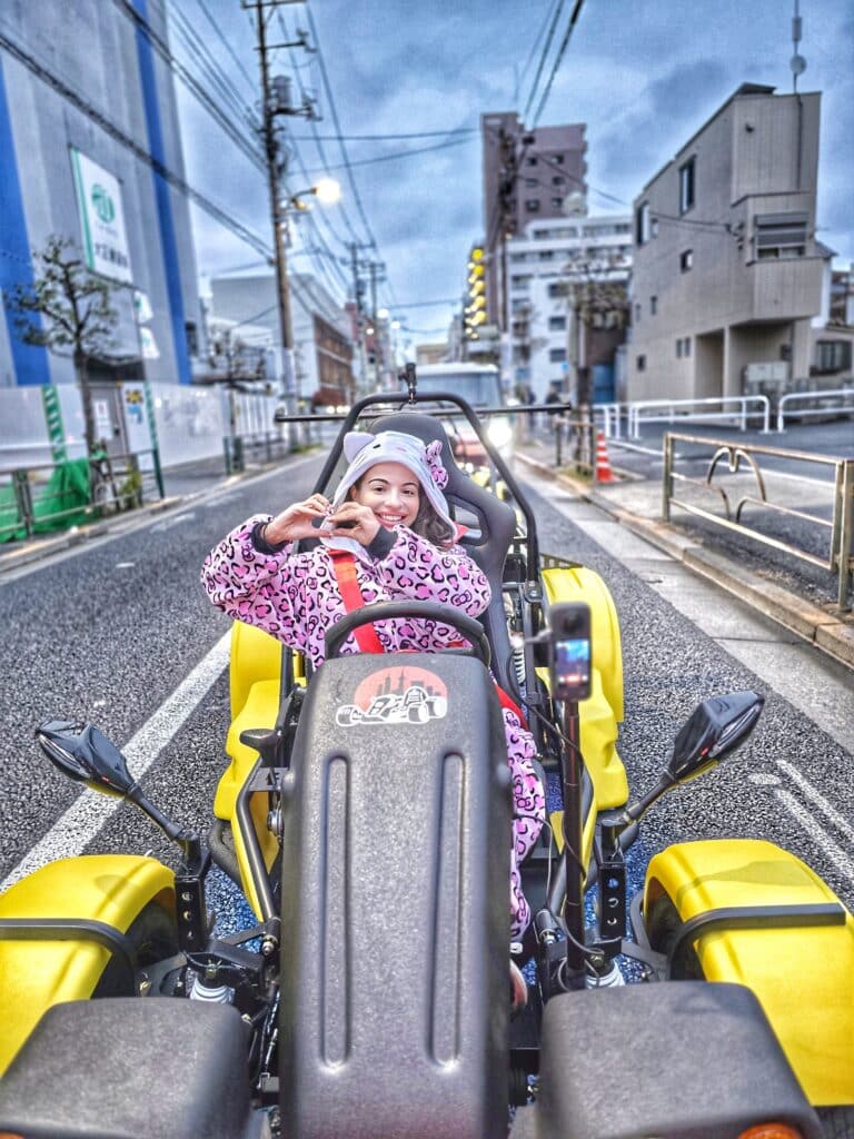 Woman in a pink leopard Hello Kitty costume makes a heart gesture while driving a go-kart on a Tokyo street with Kartzilla Go Kart.