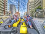 Two tourists smiling and waving while driving Mario Kart style street karts in front of Tokyo Skytree during a go-kart Tokyo tour with Kartzilla Go Kart.
