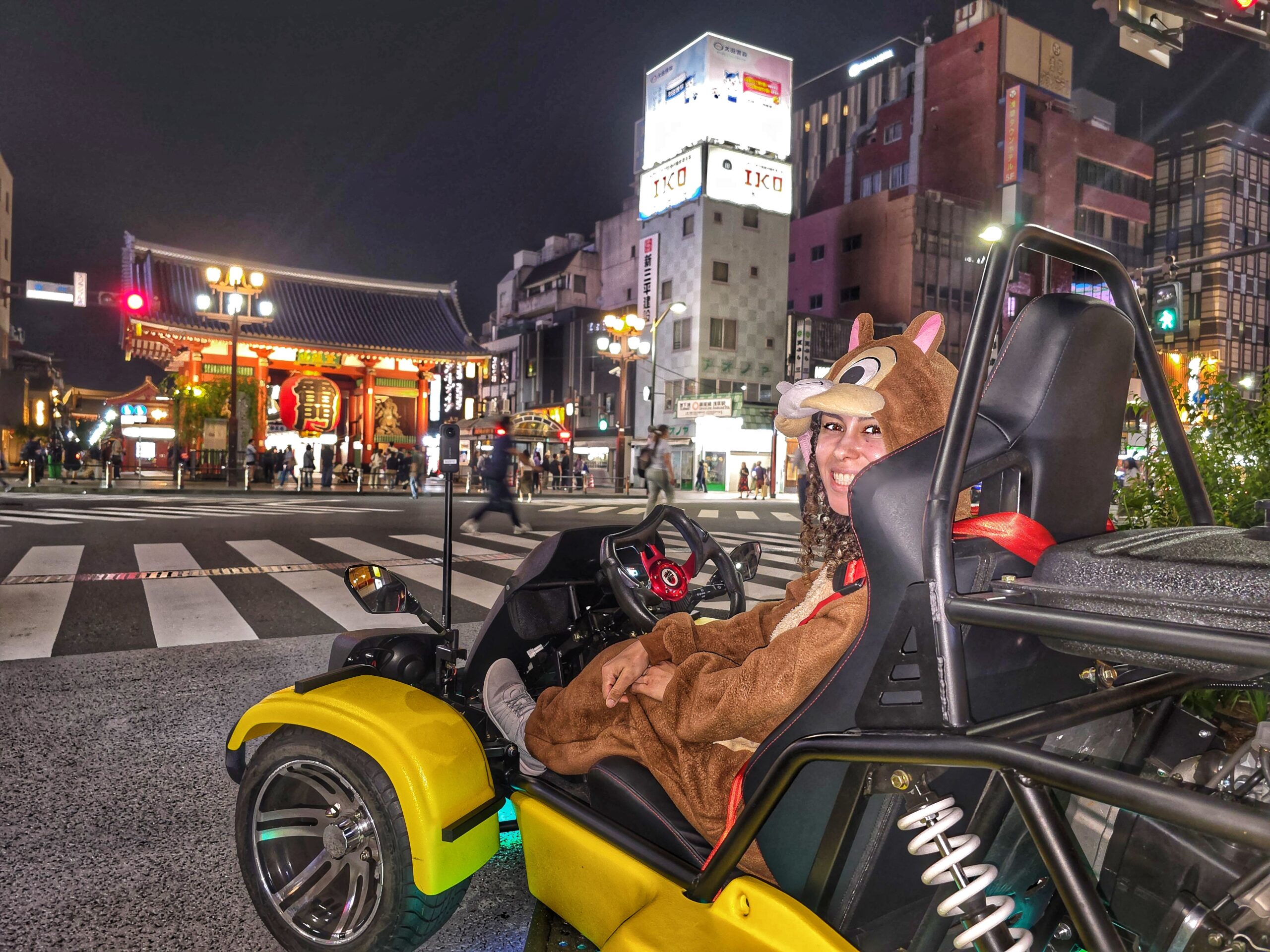 Smiling woman in a Mario Kart-style Chipmunk costume driving a yellow street go-kart near Kaminarimon Gate in Asakusa, Tokyo at night.