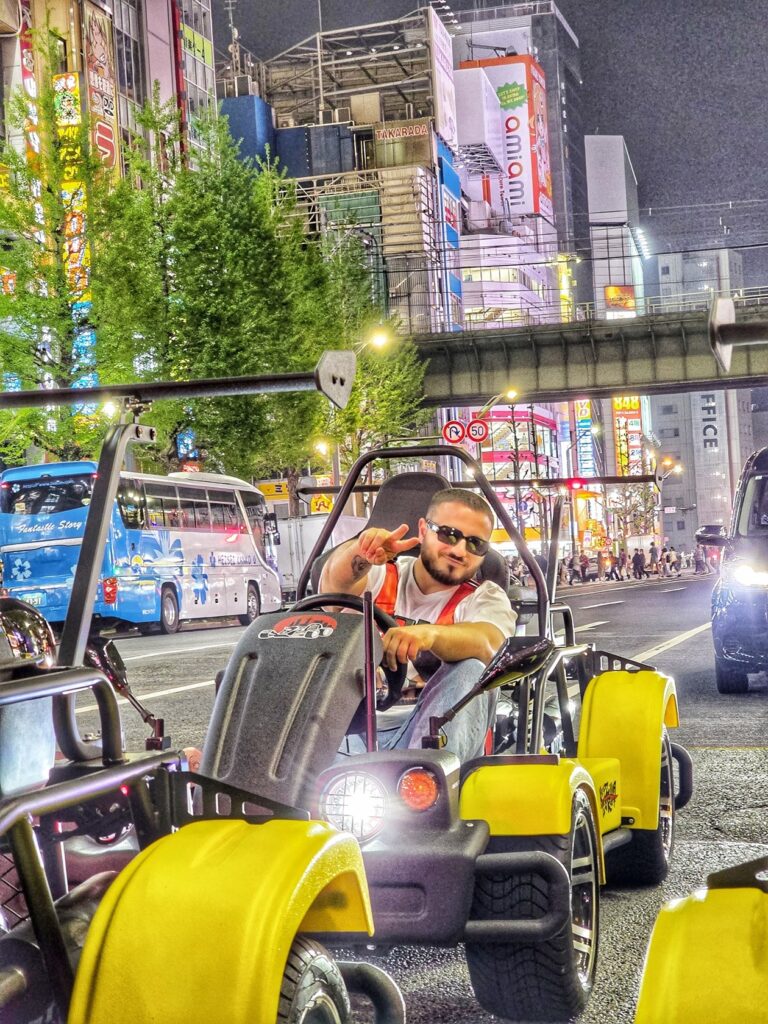 Man in sunglasses flashing a peace sign while driving a yellow go-kart through Akihabara at night during a Kartzilla Go Kart Tokyo street tour.