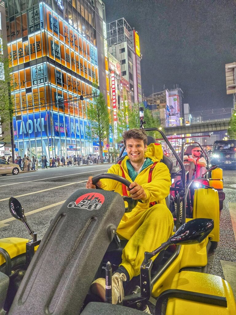 Smiling young man in a Pikachu-style costume driving a yellow Mario Kart style go-kart through Akihabara’s neon-lit streets during a night tour with Kartzilla Go Kart in Tokyo.