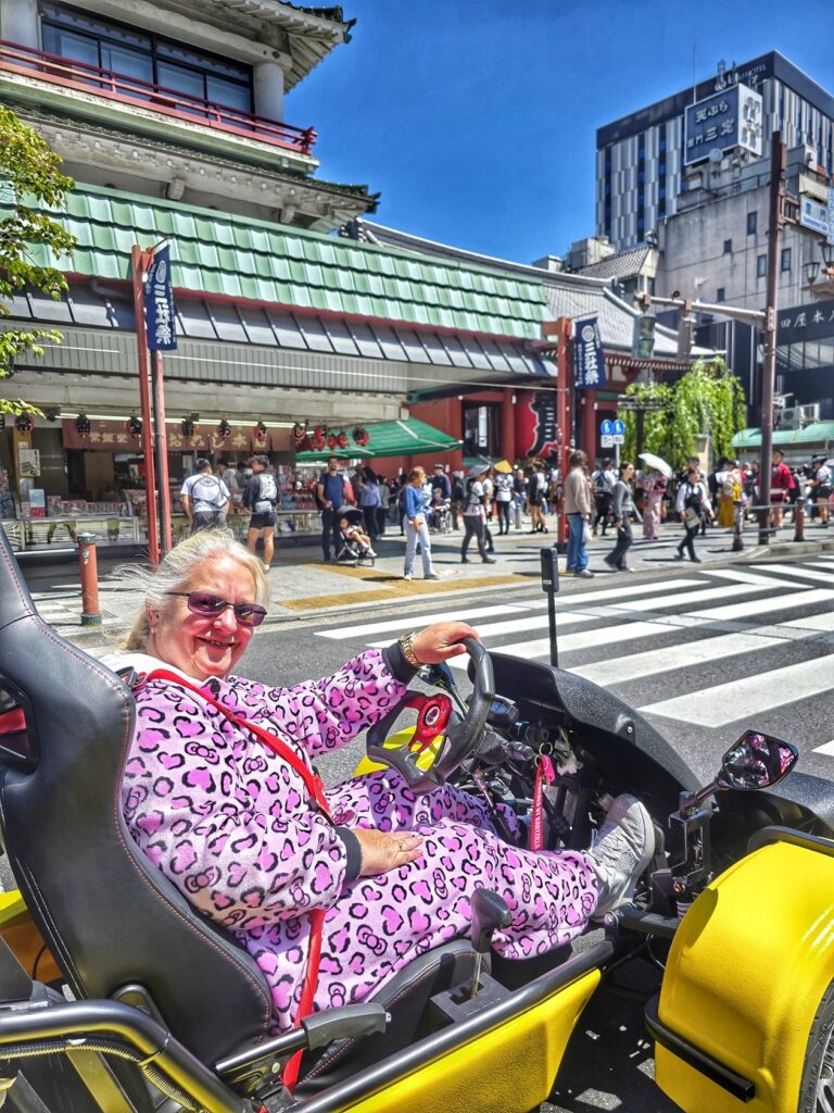 Woman in pink leopard costume driving a yellow go-kart in front of the iconic Kaminarimon gate at Asakusa during a Kartzilla Mario Kart-style tour in Tokyo.