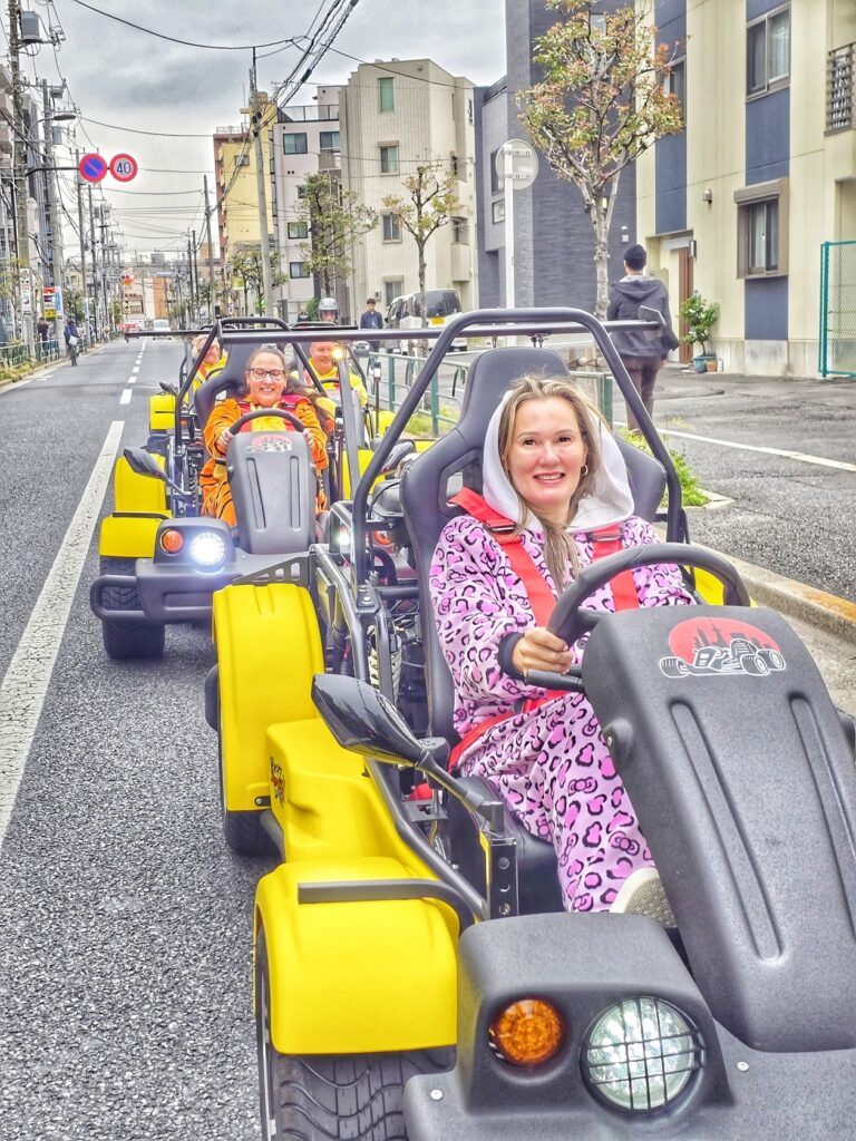 Smiling women in colorful costumes driving yellow go-karts on residential streets during a Kartzilla Mario Kart style tour in Yotsugi, Tokyo.
