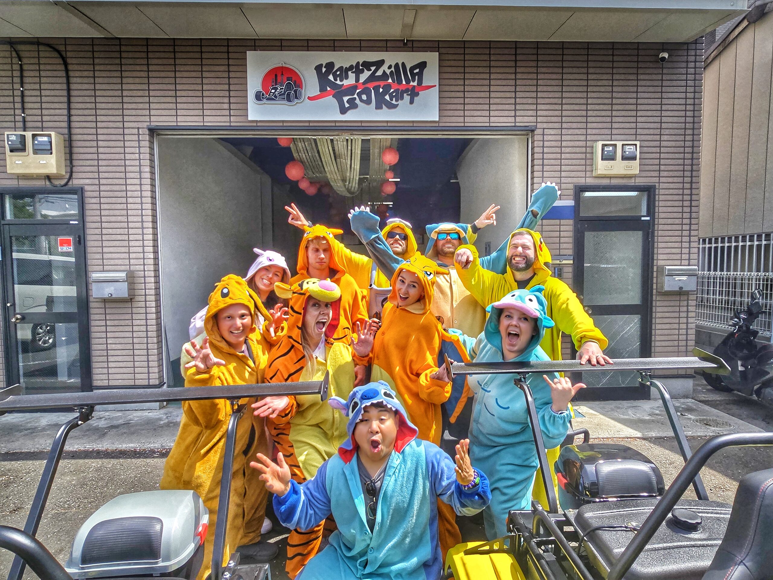 Group of tourists in colorful costumes posing excitedly in front of the Kartzilla Go Kart Tokyo facility before starting their Mario Kart style street kart tour.