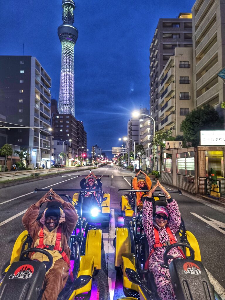 Group of people in costume driving Mario Kart style go-karts under the illuminated Tokyo Skytree at night.